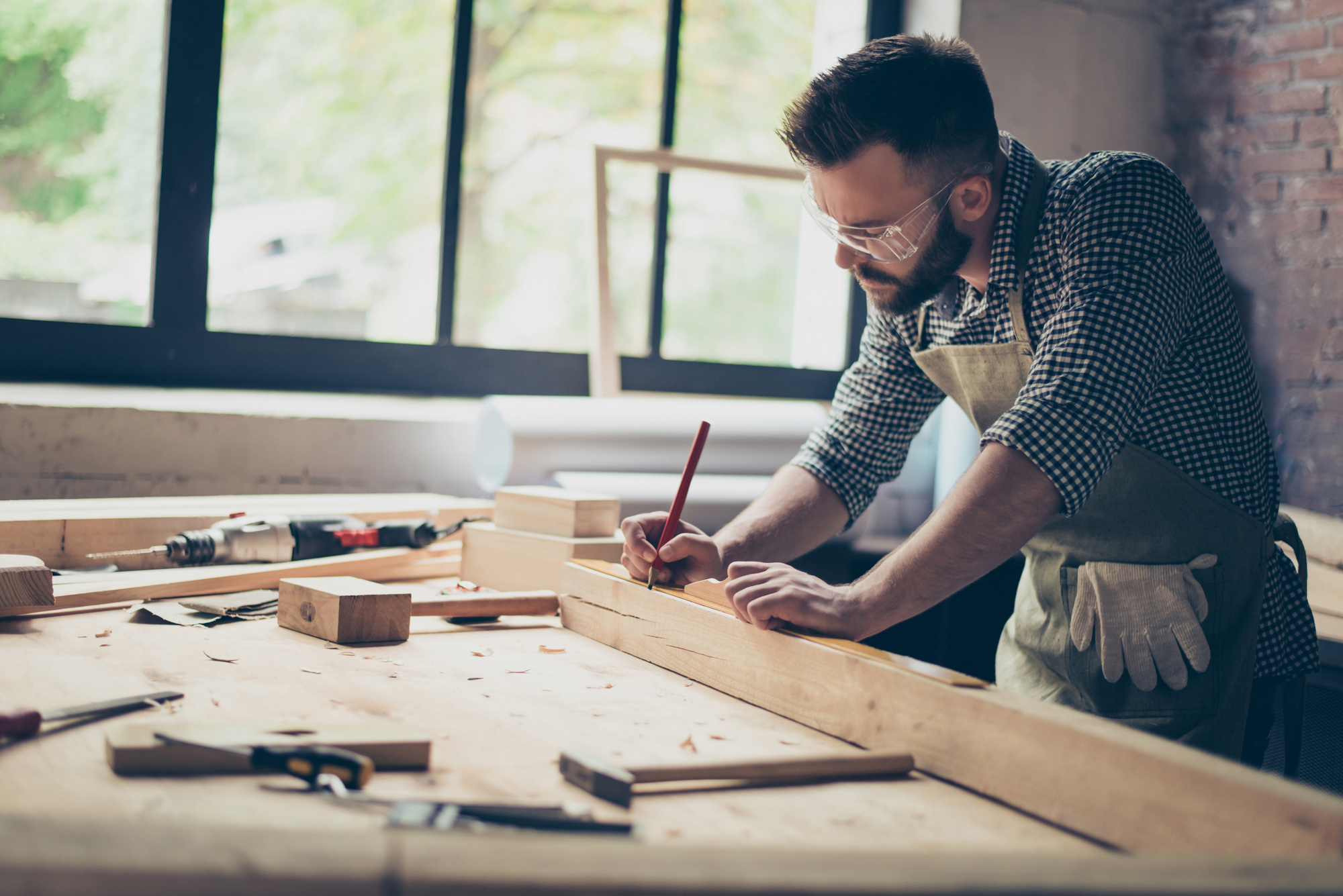 Side profile view photo of hardworking busy professional confident cabinet maker taking measure of wooden plank with a pencil near other instruments